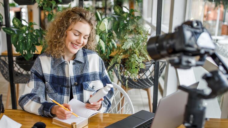 Vrouw met krullend haar leest en schrijft, camera op haar gericht in een café.
