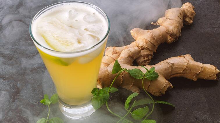 Gele drank met ijsblokjes, naast gemberwortel en groene munt.