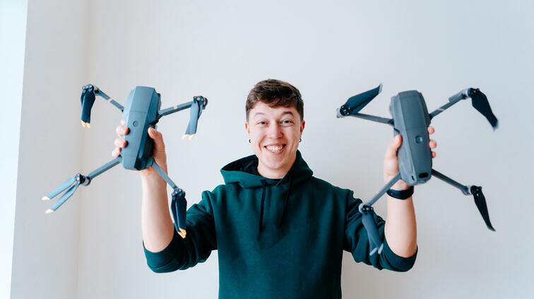 Persoon in trui houdt twee drones lachend omhoog tegen een witte achtergrond.
