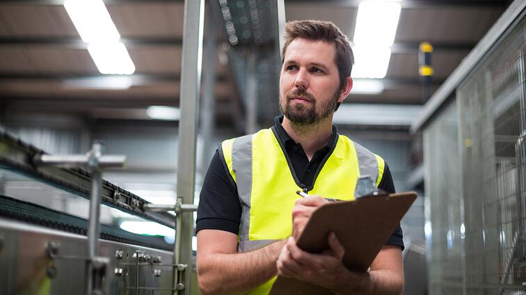 Man in gele veiligheidsvest met clipboard in fabriekshal.
