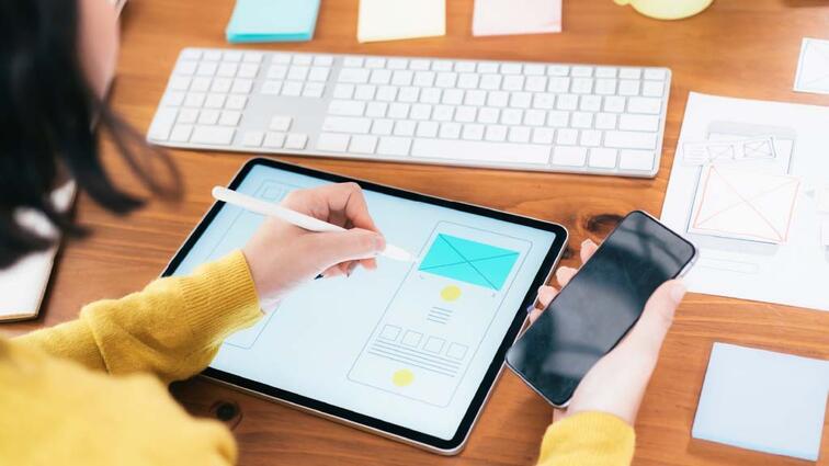 Persoon werkt op tablet met stylus, houdt smartphone vast op bureau met toetsenbord.