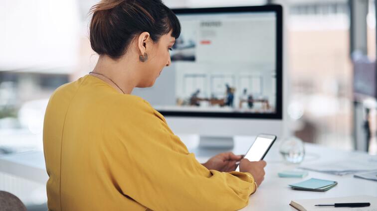Vrouw in gele trui kijkt op haar telefoon, computer op de achtergrond.