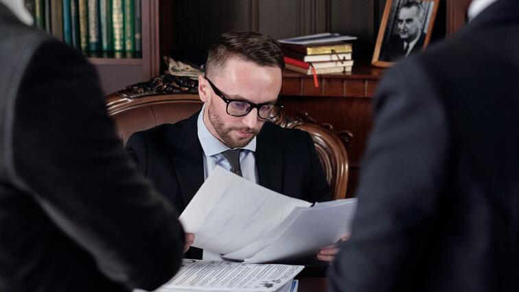 Man in pak leest documenten aan een bureau, omringd door boeken en mensen.