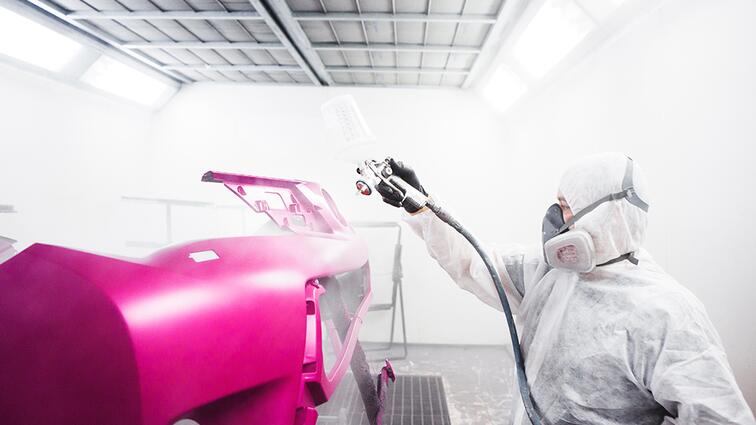 Man in witte overall spuit roze lak op auto-onderdeel in spuitcabine.