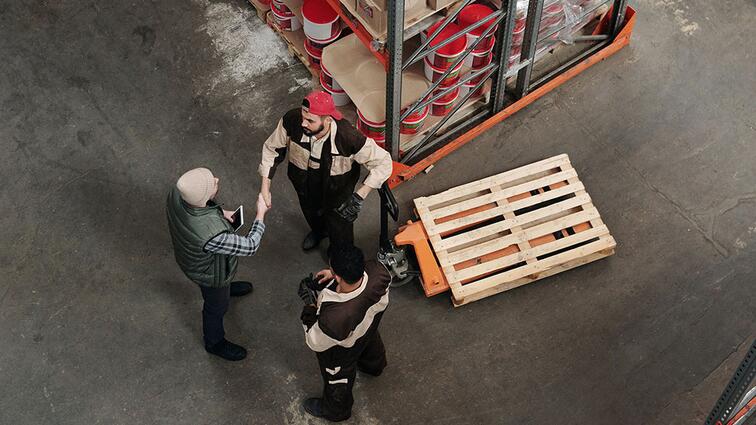 Drie mensen in een magazijn in gesprek naast een houten pallet.