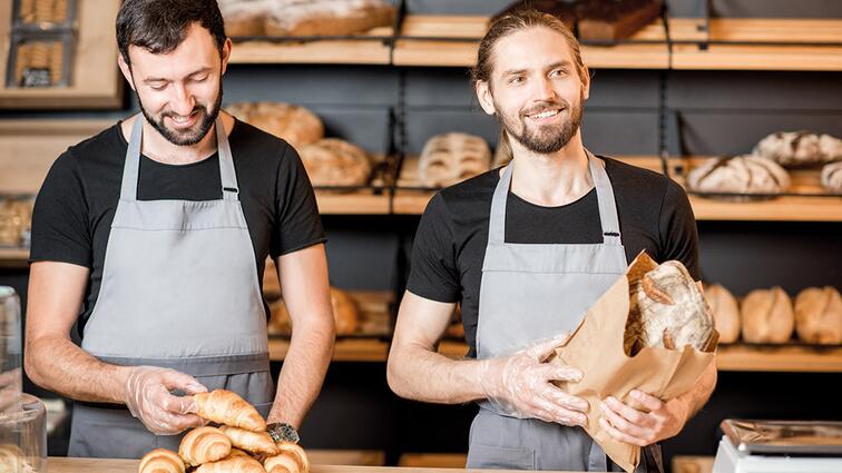 Twee bakkers in grijze schorten, lachend bij de toonbank vol broodjes.
