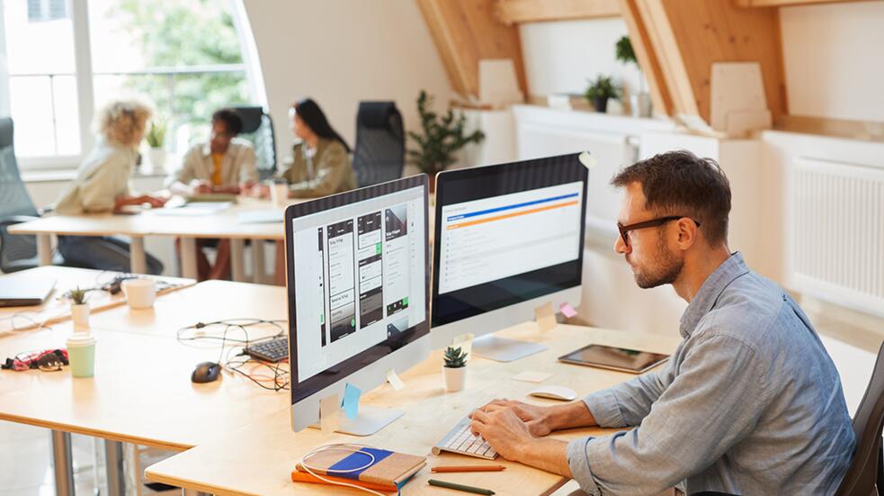 Man werkt op computer in modern kantoor, collega's overleggen op achtergrond.
