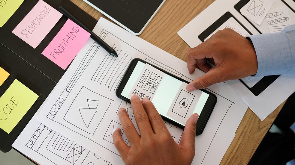 Hand houdt smartphone boven wireframe schetsen op bureau.